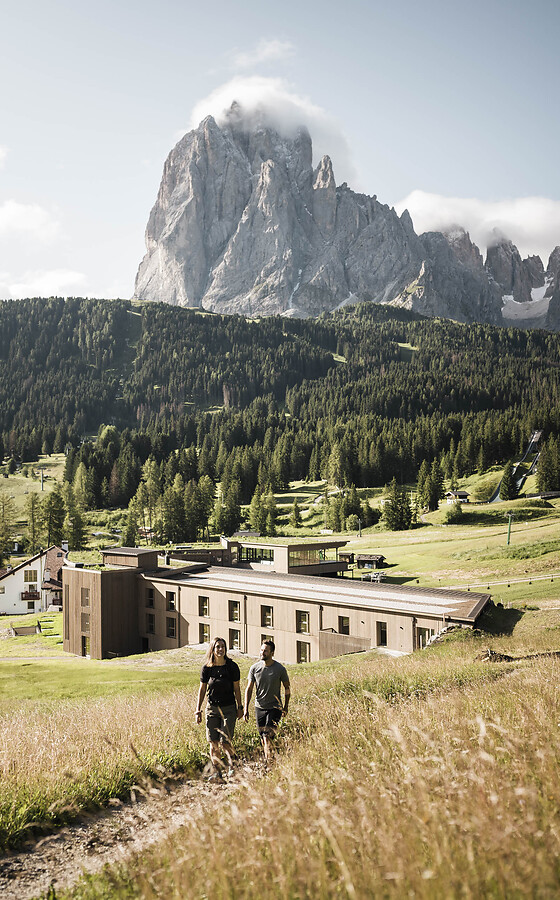 Two hikers on a meadow path in front of Hotel Cendevaves, with the striking Sassolungo in the background.